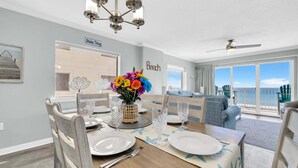 Uma área de jantar com uma mesa posta para quatro pessoas, um vaso decorativo com flores coloridas, um ventilador de teto e vista para a praia através de grandes janelas.