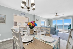 Uma área de jantar com uma mesa posta para quatro pessoas, um vaso decorativo com flores coloridas, um ventilador de teto e vista para a praia através de grandes janelas.