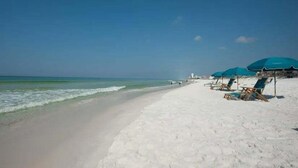 A beach with white sand, a clear blue sky, and a calm sea. There are several blue umbrellas and lounge chairs along the shore. - 🦩Book Me Baby! Take Me Away in Miramar Beach 🦩 (Miramar Beach)