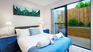 A modern bedroom with a bed, two bedside tables, a nightstand, a large window, and a sliding glass door leading to a wooden deck. - Modern Townhouse with Parking near Cornwall Park (Auckland South)