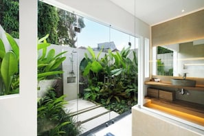 A modern bathroom with a glass shower enclosure, a sink with a large mirror, and a view of a lush garden. - Villa Aman by BaliSuperHost (Bali)