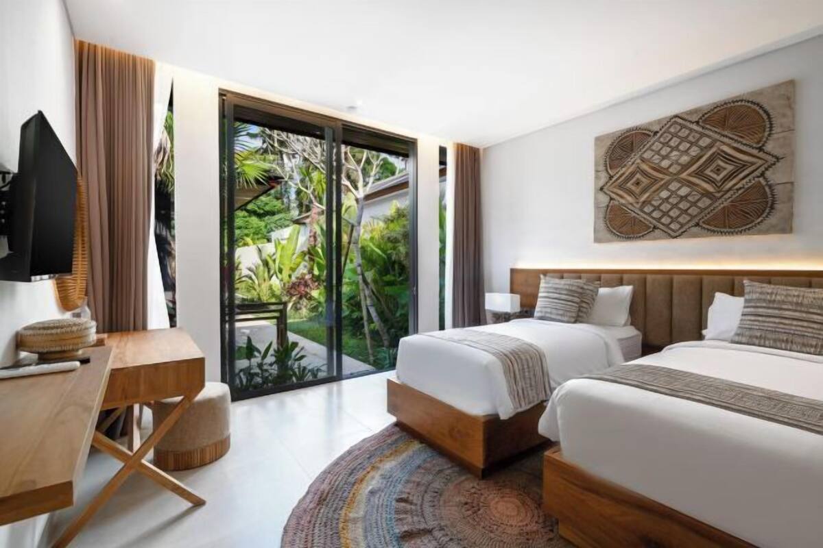 A modern hotel room with a large sliding glass door to a garden, a wooden desk, two beds, and a wall-mounted artwork.