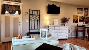 A living room with a white sofa, a black ottoman, a white cabinet, a TV mounted on the wall, and a dining area with a wooden table and chairs. - Silver Creek Accessible Studio (Helena)