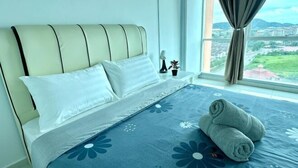 A neatly made bed with a floral bedspread and two rolled towels, a bedside table with a lamp and a small plant, and a window with a view of a cityscape and greenery. - Setia Tri-Suite(3) by Homey Planet (Bayan Lepas)