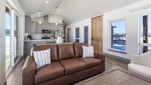 Un salón moderno con un sofá de cuero marrón, un sillón blanco y una cocina al fondo.