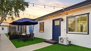 A modern house with a patio, artificial grass, a dining table with chairs, and string lights. - Designer 2BR in Prime Location w/ Outdoor Dining! (San Diego)