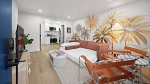 A modern living room with a sofa, a coffee table, and a TV. There is a kitchen in the background. - Designer 2BR in Prime Location w/ Outdoor Dining! (San Diego)