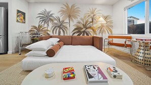 Ein Schlafzimmer mit einem Bett, Kissen, einem Nachttisch mit Büchern und einem Gesellschaftsspiel, einer Lampe und einer Wandmalerei von Palmen.