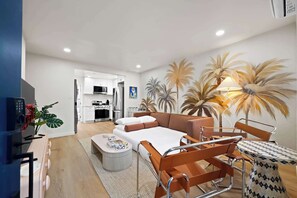 Un salon moderne avec un canapé, une table basse et une petite table avec une lampe. On aperçoit une cuisine au fond.