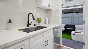 Cocina moderna con armarios blancos, un grifo negro y un lavamanos. Hay una planta en la encimera y se ve una zona de estar exterior con un sofá blanco y un hogar.