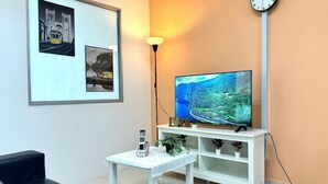 A living room with a flat-screen TV on a white stand, a small table with a phone and a plant, and framed pictures on the wall. - Setia Tri-Suite(2) by Homey Planet (Bayan Lepas)