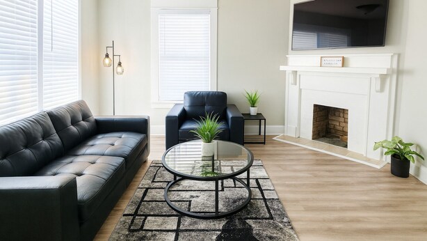 A living room with a black leather sofa, a blue armchair, a round glass coffee table, and a fireplace. - Stylish 2BR Near ECU Health, ECU & Downtown • Fast WiFi (Greenville)
