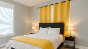 A bedroom with a bed, two bedside tables, a window with blinds, and yellow curtains. - Stylish 2BR Near ECU Health, ECU & Downtown • Fast WiFi (Greenville)