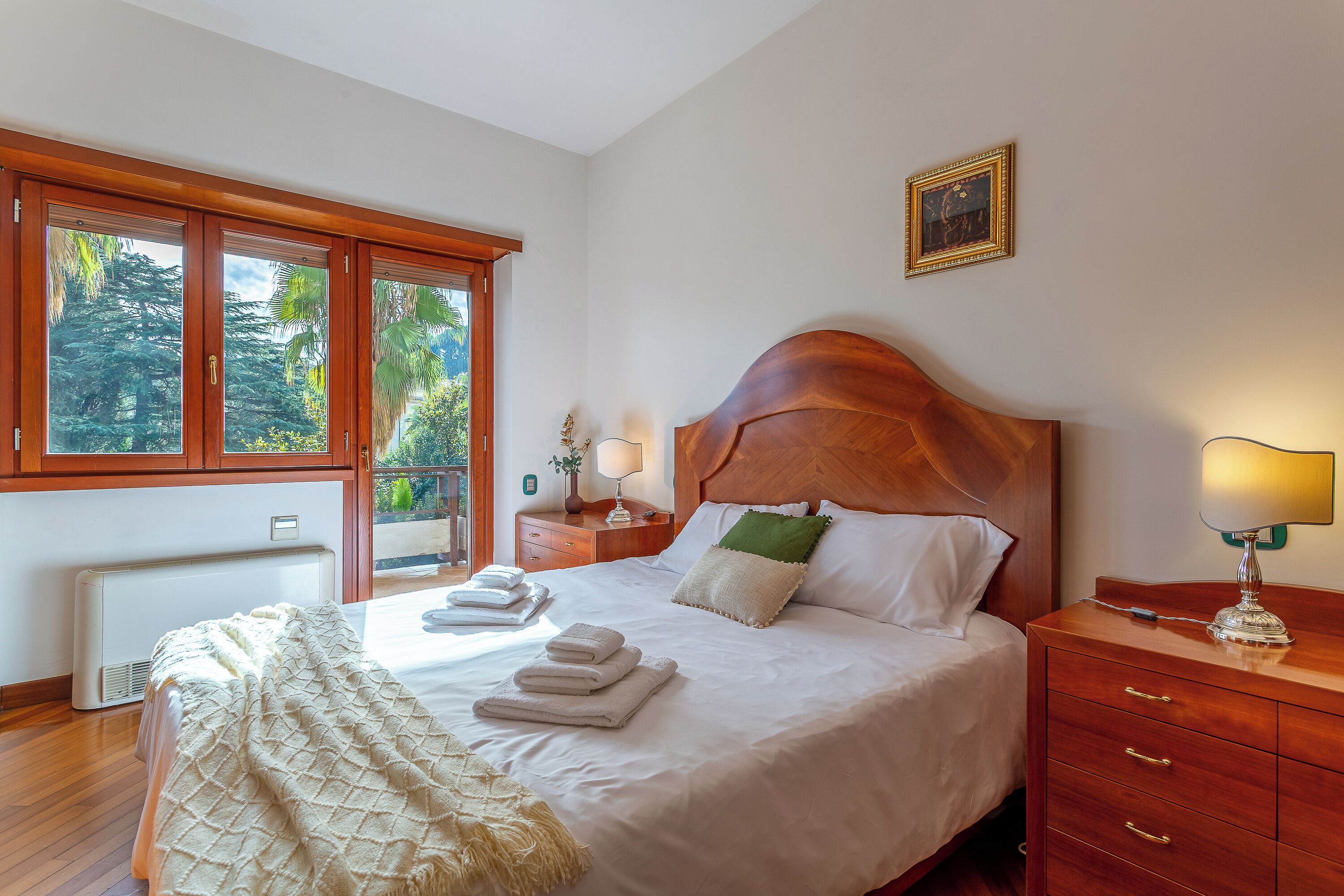 Ein ordentlich bezogenes Bett mit weißen Leinen und einem grünen Kissen, ein Nachttisch aus Holz mit einer Lampe und ein Fenster mit Blick auf Bäume.