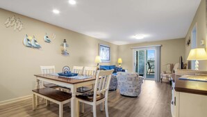 A dining area with a wooden table and white chairs, a sofa with a blue cushion, and a view through a glass door to the outside. - Harbour Club A5 (Ocean City)