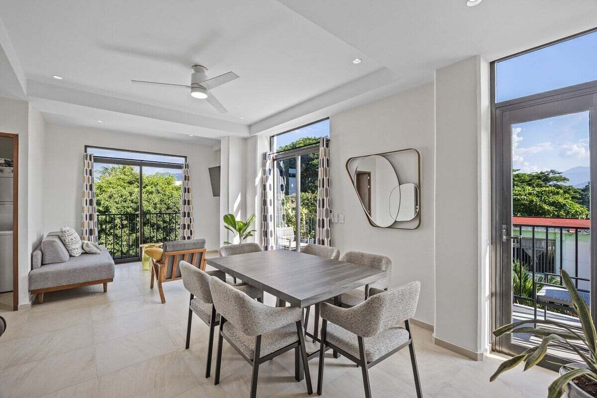 Un comedor moderno con una mesa grande y sillas, un ventilador de techo y ventanas amplias con vistas a zonas verdes y montañas.