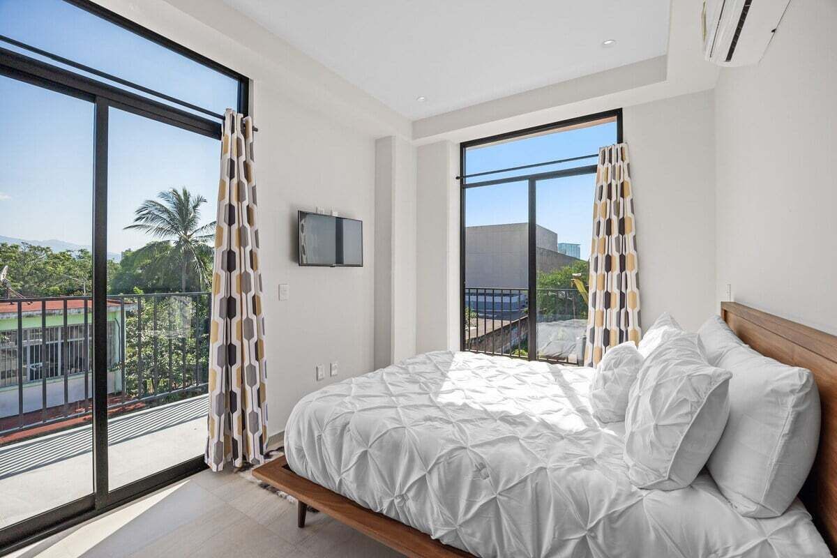 Un dormitorio moderno con una cama grande, un televisor y un balcón que ofrece vistas a palmeras y un edificio distante.