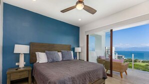Ein Schlafzimmer mit einem großen Bett, zwei Nachttischen mit Lampen, einem Deckenventilator und Meerblick durch eine Glasdoor.