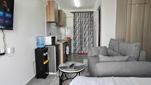 A compact living space with a grey sofa, a small round table, a water dispenser, a microwave, and a TV mounted on the wall. - Premium furnished studio in Nairobi with rooftop fitness room (Nairobi)