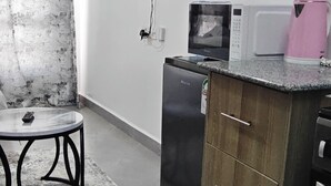 Eine kompakte Küche mit Kühlschrank, Mikrowelle und einem rosa Wasserkocher. Ein kleiner Tisch mit einer Fernbedienung und einem gemusterten Teppich.