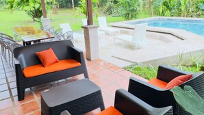 Un patio al aire libre con mobiliario de mimbre, una piscina y exuberante follaje de fondo.