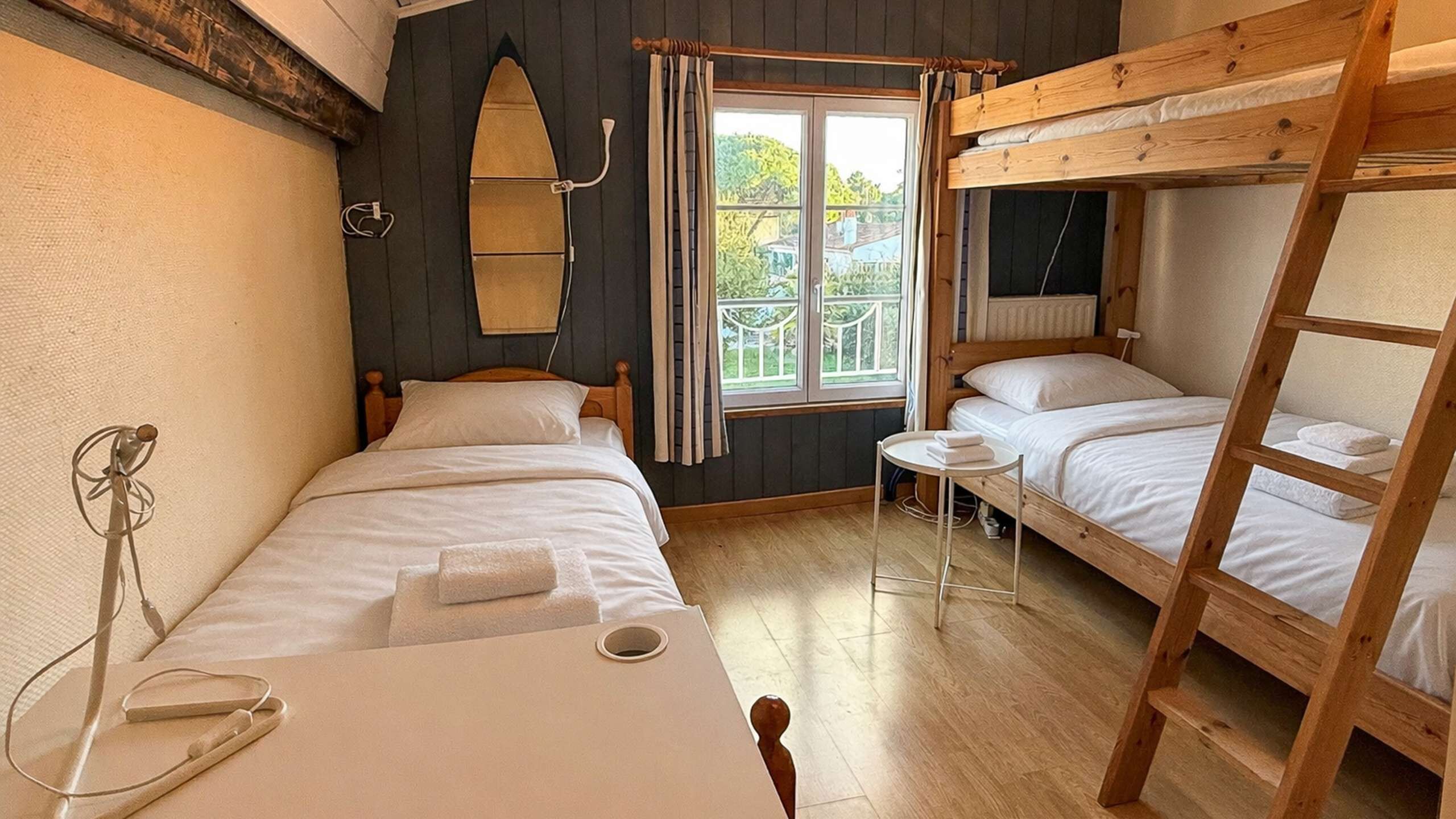 Quarto com beliches, uma mesinha com uma toalha, uma cadeira e janela com cortinas.