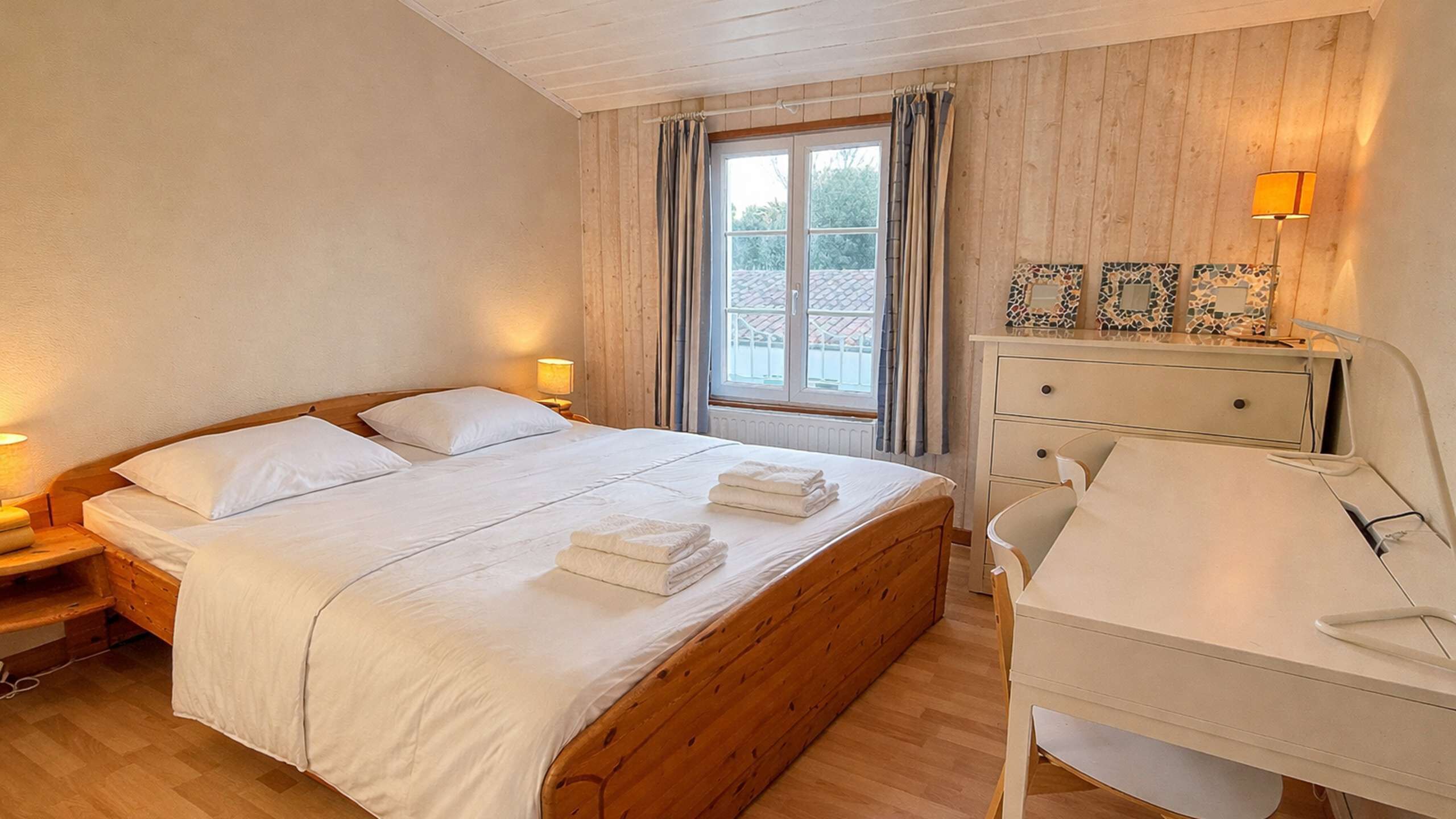 A bedroom with a wooden bed, a bedside table with a lamp, a window with curtains, and a desk with a chair.
