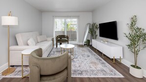 Ein modernes Wohnzimmer mit einer Couch, einem Sessel, einem Couchtisch, einem Fernseher und einem großen Fenster mit Jalousien.