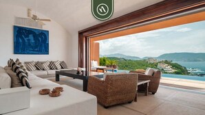 A modern living room with a sofa, coffee table, and wicker chairs, overlooking a scenic view of a coastline and mountains. - 4 BR Penthouse - Breathtaking Views - Private Pool (Zihuatanejo)
