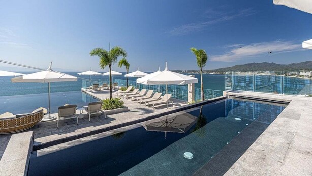 Una piscina infinita con sillones y sombrillas, con vistas a una masa de agua y montañas distantes.