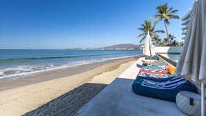 Un salón junto a la playa con vista al océano, palmeras y una montaña al fondo.