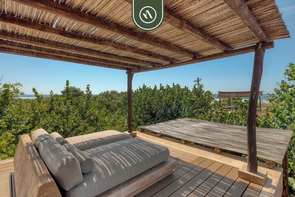 A wooden deck with a cushioned seating area, a thatched roof, and a view of the ocean.