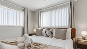 Ein ordentlich bezogenes Bett mit beiger Decke und Kissen, ein Holzkopfteil und ein Nachttischchen mit Lampe.
