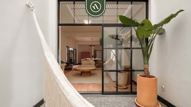 Un couloir avec un hamac, une grande plante en pot et une porte-fenêtre coulissante donnant sur un espace de vie comprenant une table ronde et des chaises.