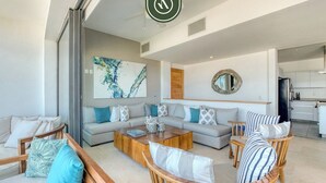 Un salon moderne avec un ensemble de canapés, une table basse et des chaises en bois. Un grand tableau abstrait et un miroir ornent les murs.