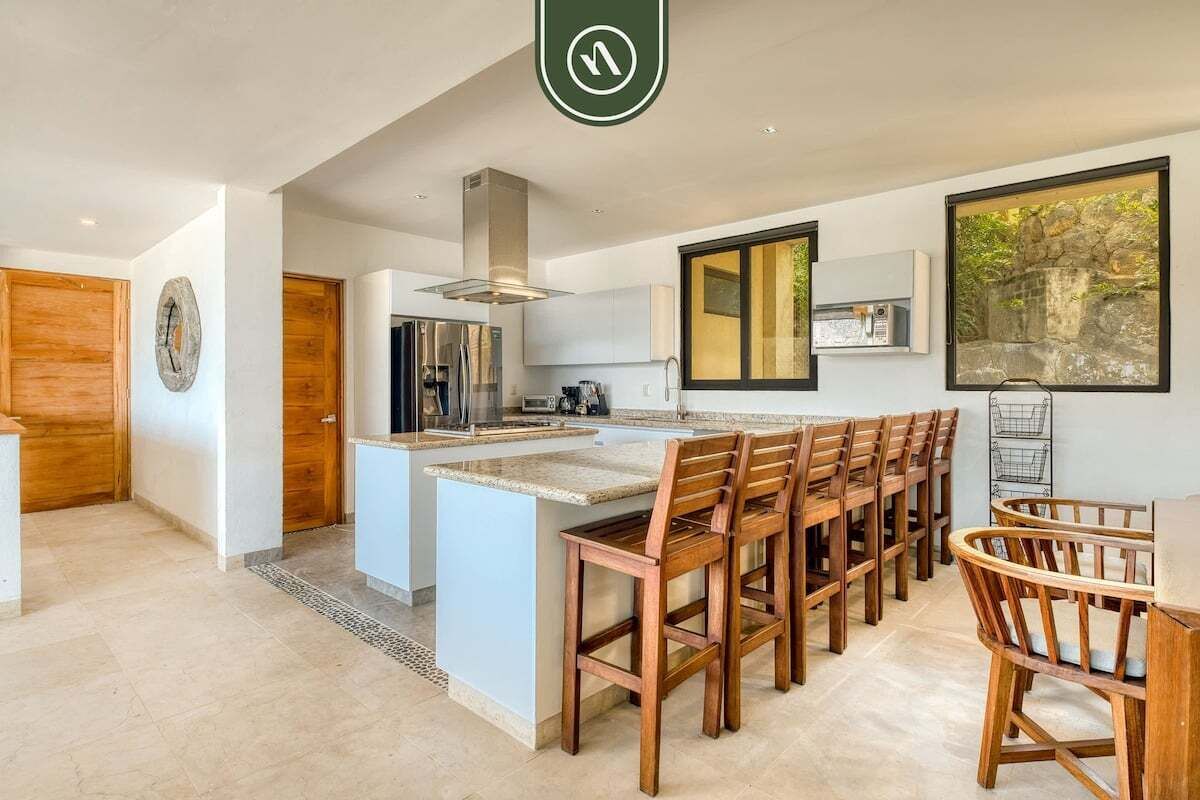 A modern kitchen with a central island, wooden chairs, and a large window with a view of greenery.