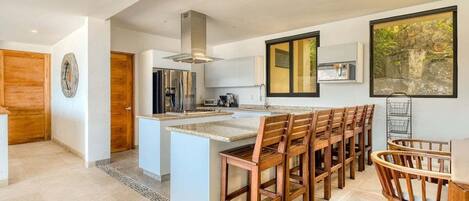 A modern kitchen with a central island, wooden chairs, and a large window with a view of greenery.