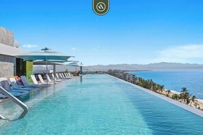 Una piscina con sillas de descanso y sombrillas, con vista a una playa y montañas.