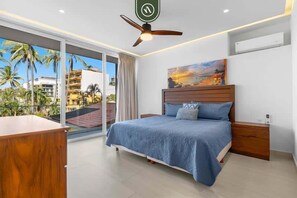 Un dormitorio con cama, escritorio, ventilador de techo y una ventana grande con vista a palmeras y edificios.