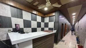 A reception area with a white counter, black and white checkered wall panels, a ceiling fan, and a hallway with potted plants. - Hotel Natraj A Boutique Hotel (Haridwar)