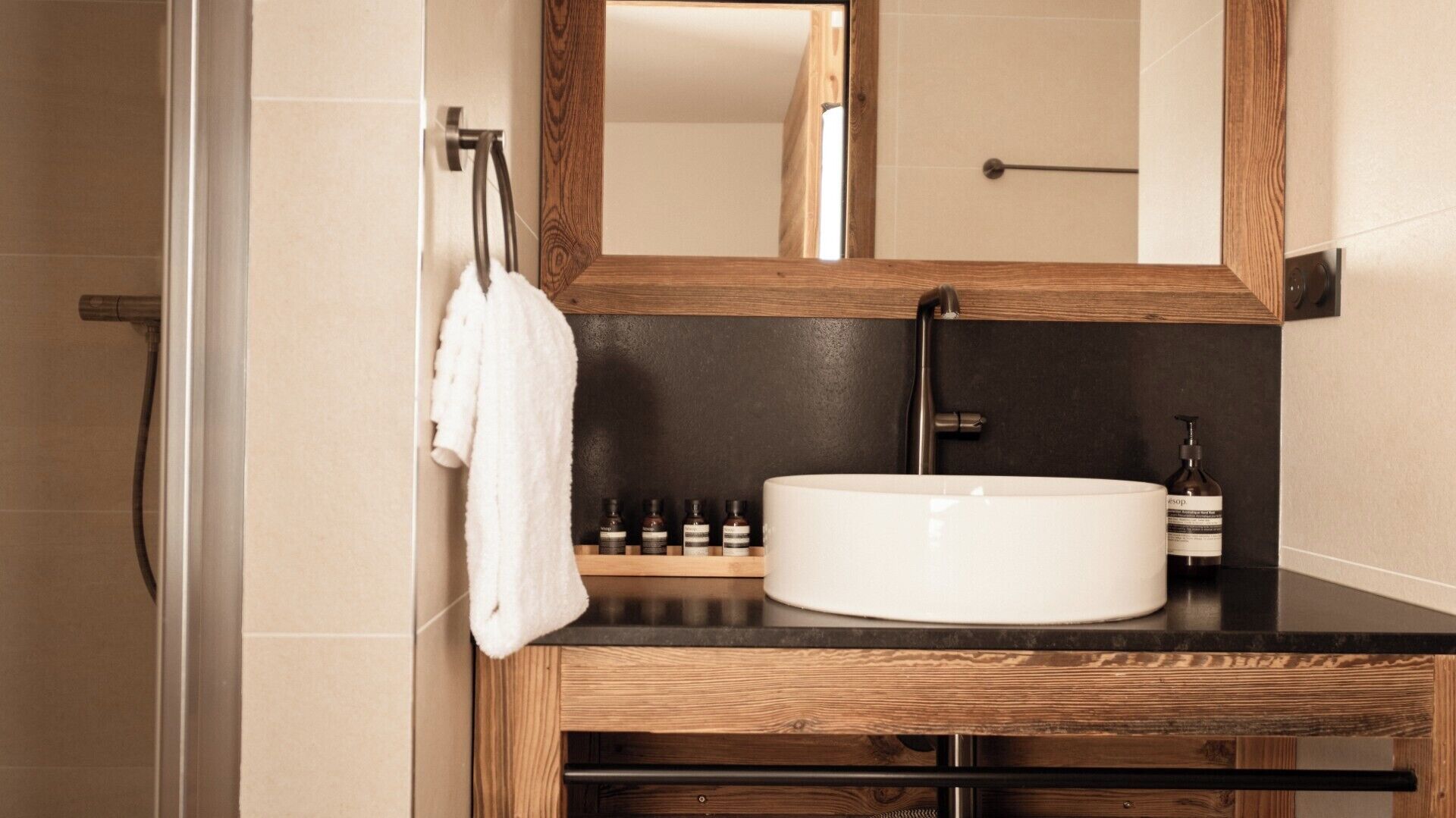 Ein Badezimmer mit einem Holzlagerfläschchen, einem weißen Waschbecken und einem Spiegel. Auf dem Lagerfläschchen stehen Pflegeprodukte und ein Handtuch hängt an einem Haken.