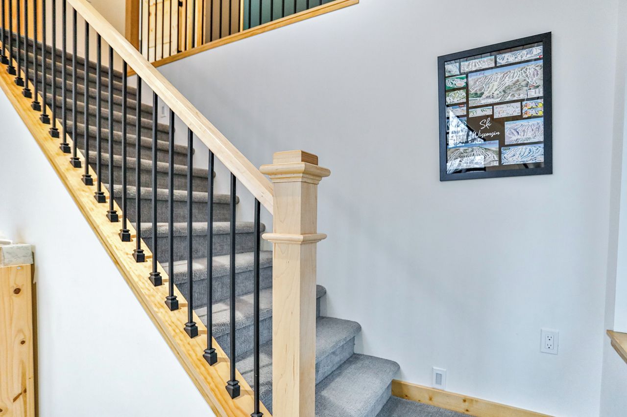 Eine Treppe mit hölzernen Geländern und schwarzen Spindeln, die zu einer Zwischenebene mit einer gerahmten Bildercollage an der Wand führt.
