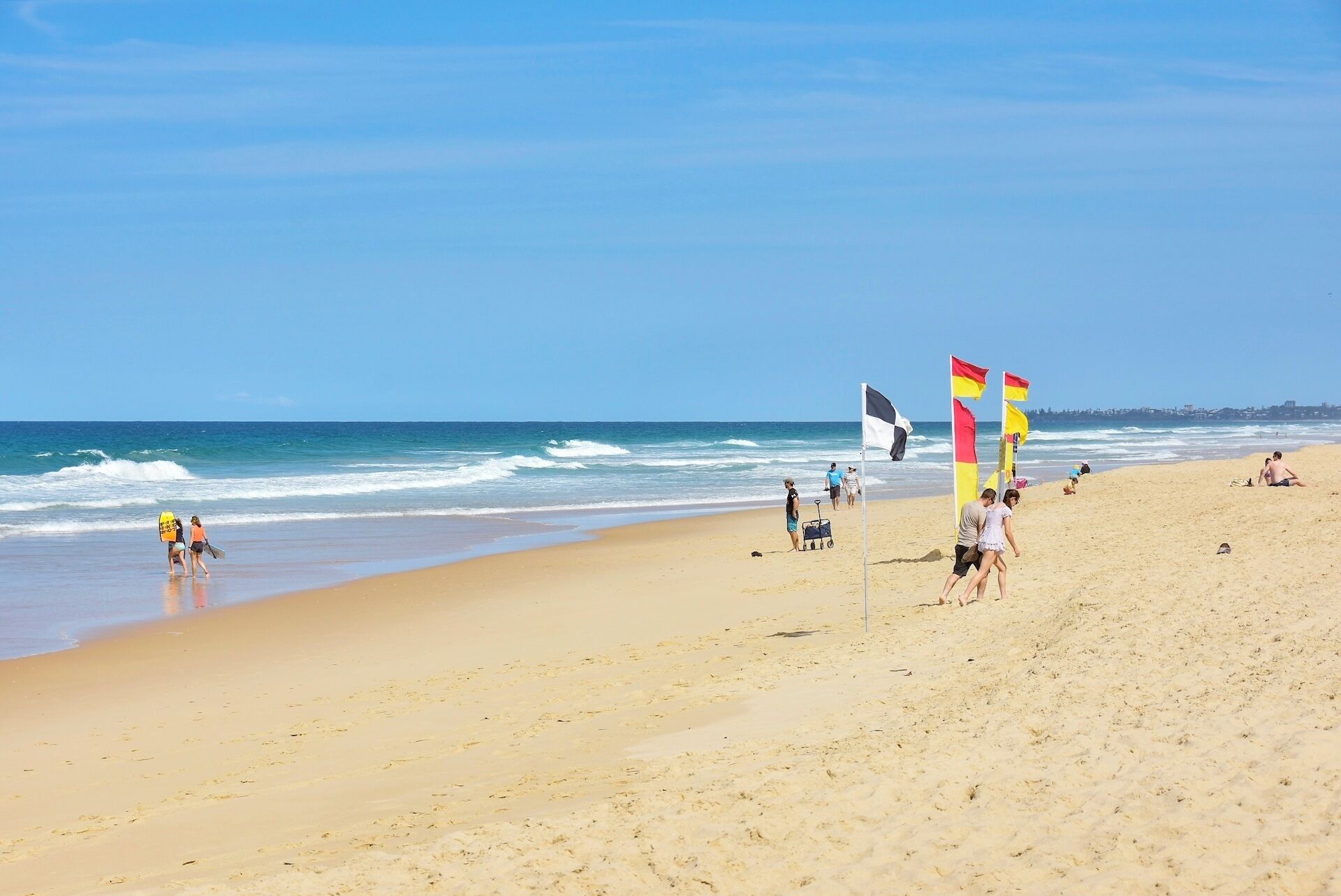 A beach with people walking, a lifeguard flag, and a clear sky.