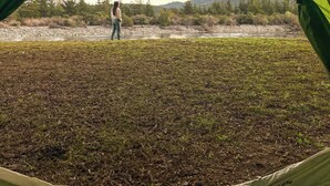 A tent with a person standing inside, overlooking a natural landscape with trees and a body of water. - NaboCamp (Linares)