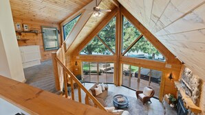 A spacious loft-style room with a high ceiling, large windows, a fireplace, and a cozy seating area. - Risa Cabin: Full Lodge | Lake, Hot Tub, 9 Acres (Bend)
