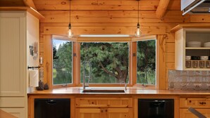A wooden kitchen with a built-in dishwasher, a window with a view of trees, and hanging light fixtures. - Risa Cabin: Full Lodge | Lake, Hot Tub, 9 Acres (Bend)