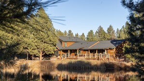 A log cabin with a porch, surrounded by trees and a pond. - Risa Cabin: Full Lodge | Lake, Hot Tub, 9 Acres (Bend)