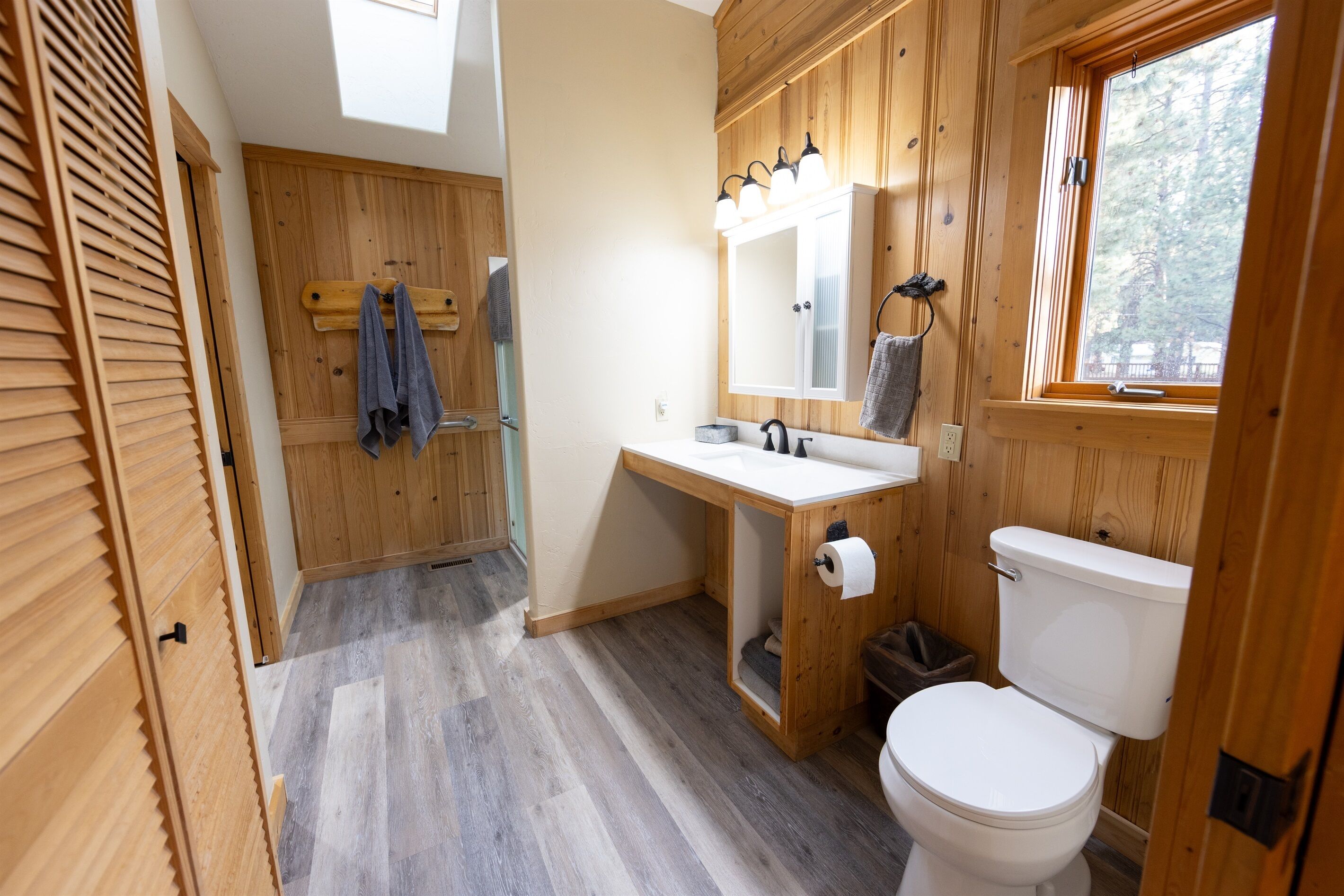 A bathroom with wooden walls, a white sink, a toilet, and a window with a view of trees.