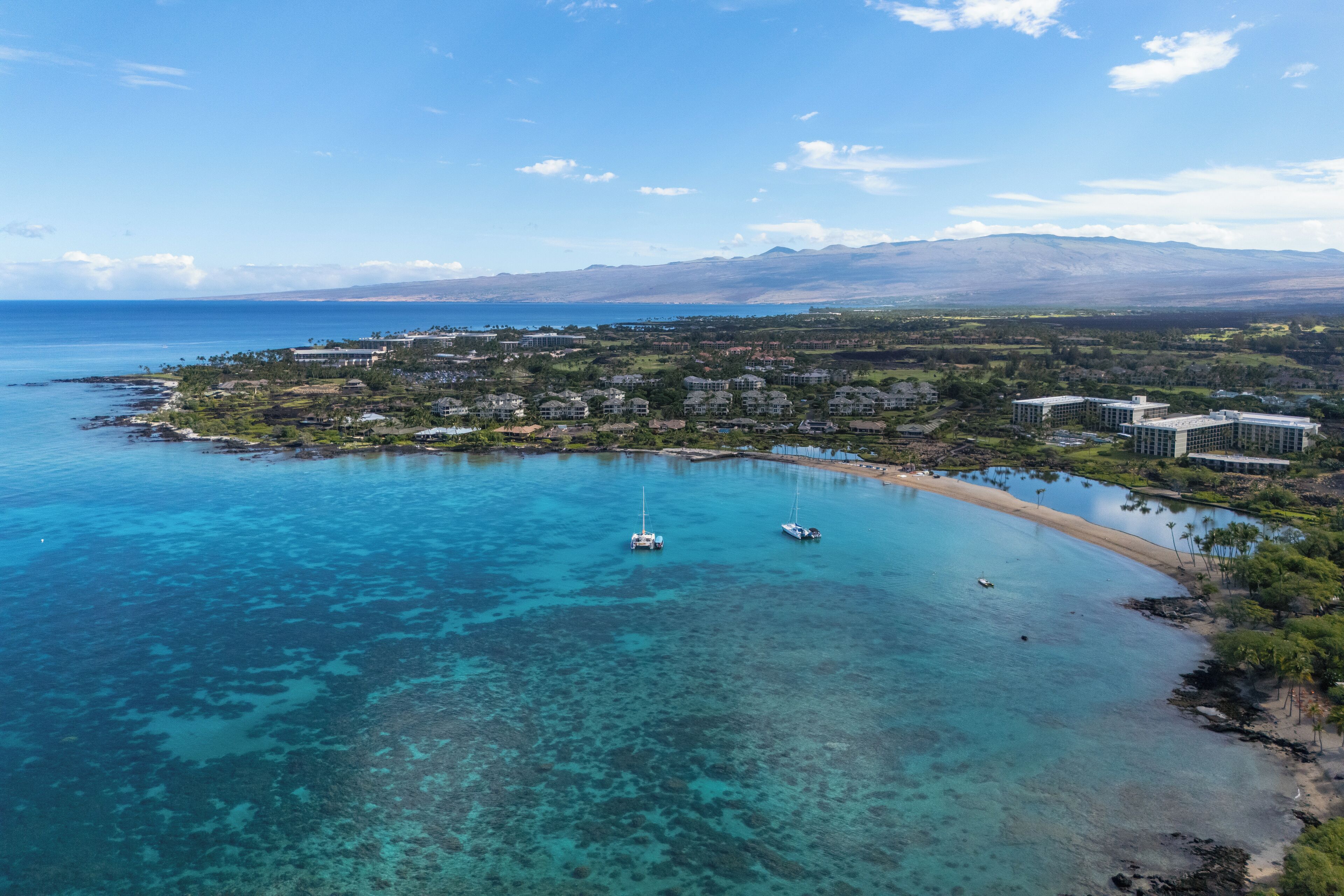Vista aérea de un complejo turístico en la costa, con aguas turquesas cristalinas, una playa de arena y montañas al fondo.