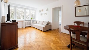 A spacious living room with a herringbone wood floor, a white sofa, a wooden dining table with chairs, and a television on a wooden cabinet. - Mokotów Urban Comfort Apartment (Warszawa)